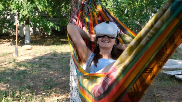 Радостная женщина в очках виртуальной реальности лежит в гамаке рядом с деревом, представляя, как сбывается мечта, выдуманный мир - Фото, изображение