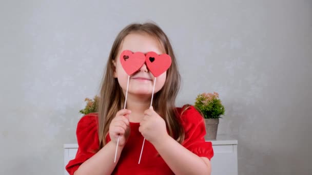 Mooi klein meisje in een rode jurk met twee harten op een stok sluit haar ogen met hen. Valentijnsdag concept. - Video