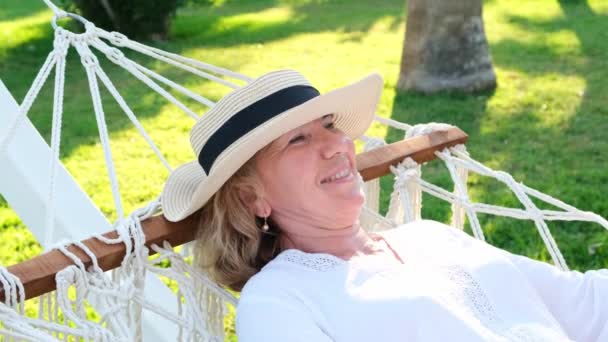 mujer anciana en un sombrero de paja está descansando en una hamaca alrededor de las palmeras y disfrutando de la tranquilidad y la relajación. Concepto de estilo de vida - Imágenes, Vídeo