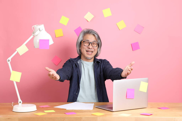 The senior Asian man sitting at working desk with a lot of paper work against the pink background. - Foto, afbeelding