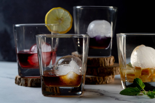 meervoudige whiskey glazen gevuld met verschillende gekleurde geïnfundeerde glühwein gin rum wodka dranken met ijsballen en schijfjes limoen, munt toont de hobby van gin infusie thuis als een viering drankje - Foto, afbeelding