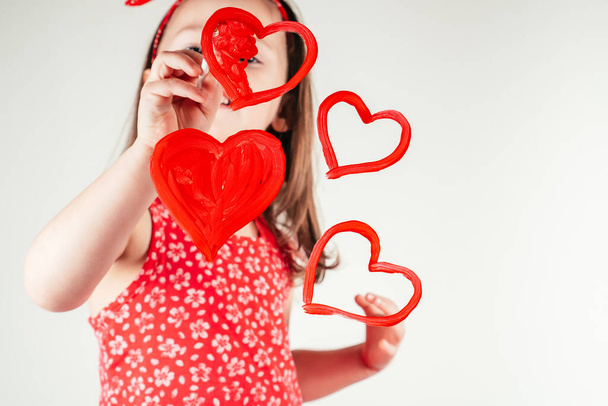 Маленькая девочка с длинными вьющимися волосами в красной одежде рисует сердца с краской на прозрачном стекле улыбаясь на белом фоне. - Фото, изображение