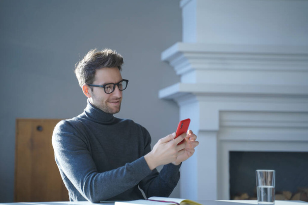 Joven hombre alemán guapo en gafas sentado en la mesa y el uso de teléfono móvil en casa - Foto, Imagen