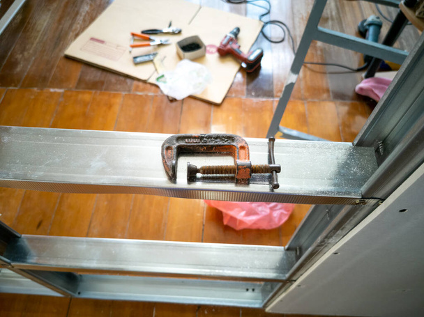 High angle view, C-clamp, placed on Galvanized frame. Blurred back, wooden floor and drill, plier, screw, bolt, grinding machine. - Foto, Imagem