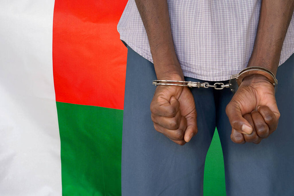Αφρικανός με χειροπέδες στο φόντο της σημαίας της Μαδαγασκάρης. Πίσω όψη. Έννοια των φυλακών και της διαφθοράς στη Μαδαγασκάρη - Φωτογραφία, εικόνα