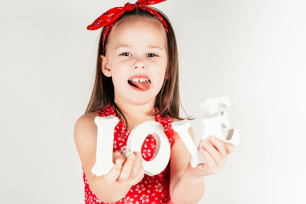 Kis aranyos lány piros ruhában tartsa fehér volumetrikus betűk szó szerelem a kezében mutatja nyelv fehér háttér. - Fotó, kép