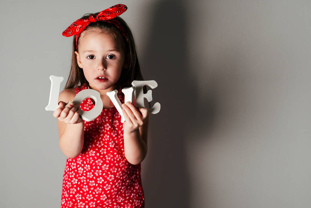 Linda niña con ropa roja sostiene letras volumétricas blancas de la palabra amor en las manos sobre fondo gris con sombra. - Foto, imagen