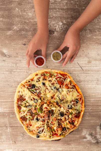 close-up van persoonlijke handen bereiden tafel met fast food, pizza met kaas, olijven, ham en peperoni, met potten van kruiden, lifestyle en restaurant menu, houten textuur achtergrond - Foto, afbeelding