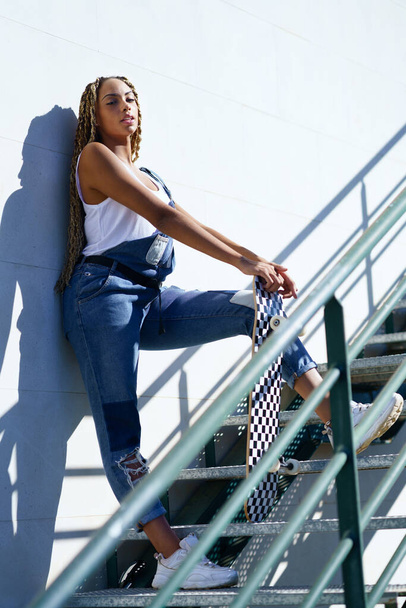 Молода чорна дівчина з афро косами на міських сходах, що несе скейтборд
. - Фото, зображення