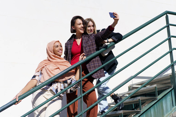 Cheerful multiracial girlfriends taking selfie on smartphone on stairs - Photo, Image