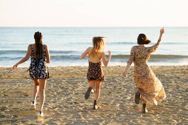 Φιλενάδες με καλοκαιρινά φορέματα τρέχουν προς την κυματιστή θάλασσα - Φωτογραφία, εικόνα