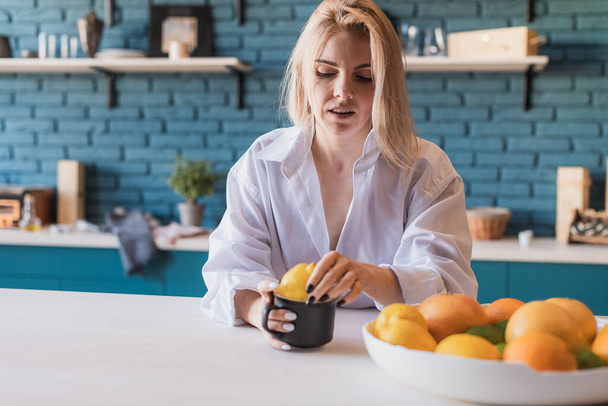 pretty, cheerful girl with blond hair, holding a cup in which yellow lemons, vitamins, in a white men's shirt, enjoys a cozy quiet morning, tastes good. - Photo, Image
