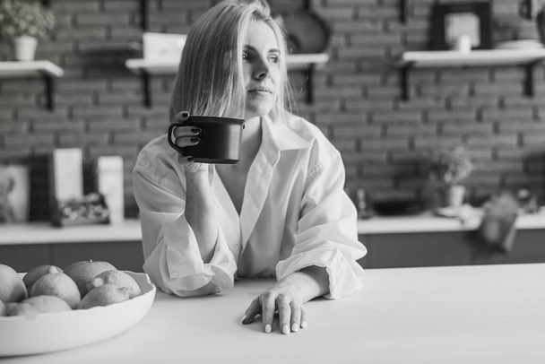 Ντροπαλή νεαρή γυναίκα κρύβεται πίσω από μια κούπα καφέ, κρατώντας την μπροστά από το πρόσωπό της με τα δύο χέρια, σε ένα λευκό ανδρικό πουκάμισο, ηλιόλουστο πρωινό, ρομαντικό - Φωτογραφία, εικόνα