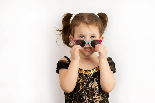 Mała radosna dziewczynka z dwoma końskimi ogonami i czarno-złotą sukienką, uśmiechnięta, zdejmuje swoje piękne okulary. Studio zdjęcie izolowane na białym tle - Zdjęcie, obraz