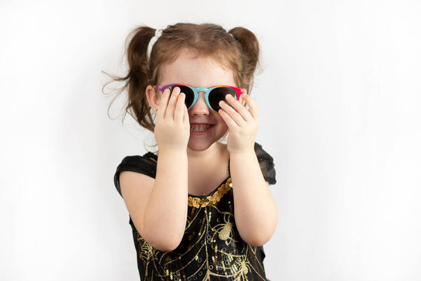 Маленькая девочка с двумя хвостиками и черно-золотым платьем закрывает свои солнечные очки руками. Студийное фото ребенка на белом фоне - Фото, изображение