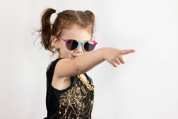 Little cheerful girl with two ponytails and a black and gold dress in sunglasses. Studio photo on a white background of a sad child pointing with his index finger - Fotó, kép