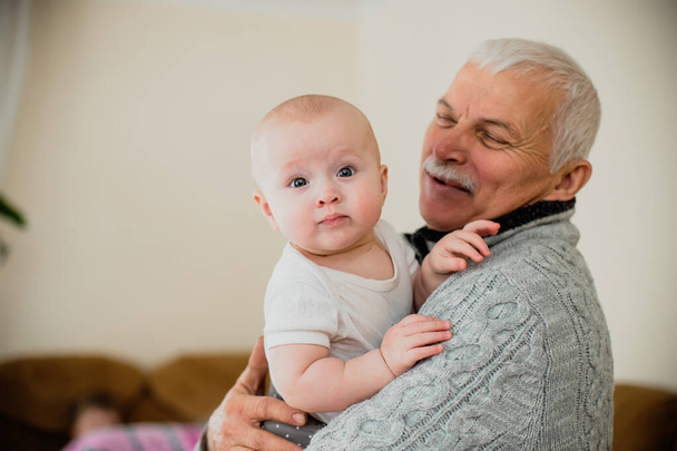 Улыбающийся старик держит ребенка на руках. Пожилой человек держит малыша в светлой комнате. Пожилой человек играет с ребенком. - Фото, изображение