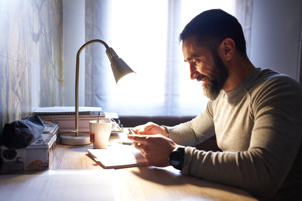 Young white man with a neat beard studying and looking at the phone at his desk - Foto, imagen