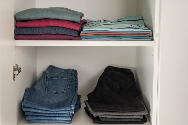 Neatly folded sweaters and jeans and other clothes on shelves in wardrobe - Photo, image