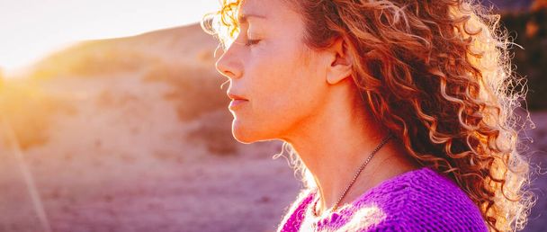 Close up sunset portrait of attractive woman with closed eyes and sun in back light. Dreaming and enjoying feeling concept lifestyle emotion. Serene female people outdoor with curly hair - Foto, Imagem
