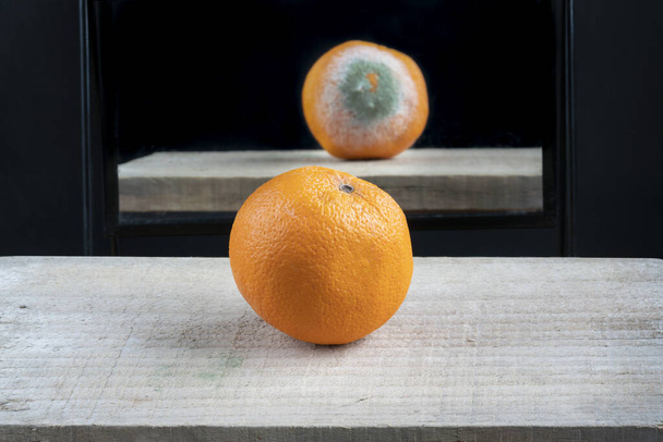 az igazi arcát látni valaminek a tükörben. Friss és érett narancs gyümölcs az asztalon a tükörreflexió ellen, rothadt rossz oldallal. Szelektív fókuszú gyümölcs. - Fotó, kép