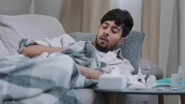 Sick health man arabic spaniard guy feeling symptoms of coronavirus covid coughing flu suffering from illness infectious virus lying on couch under blanket at home looking at table with medications - Footage, Video