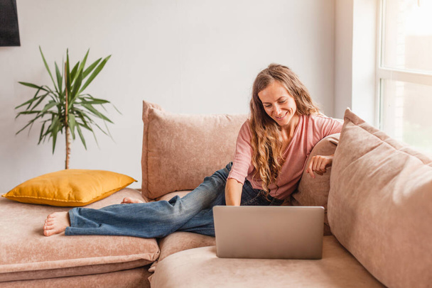 Beautiful cheerful woman using silver laptop while sitting on sofa in living room at home - Foto, Imagem