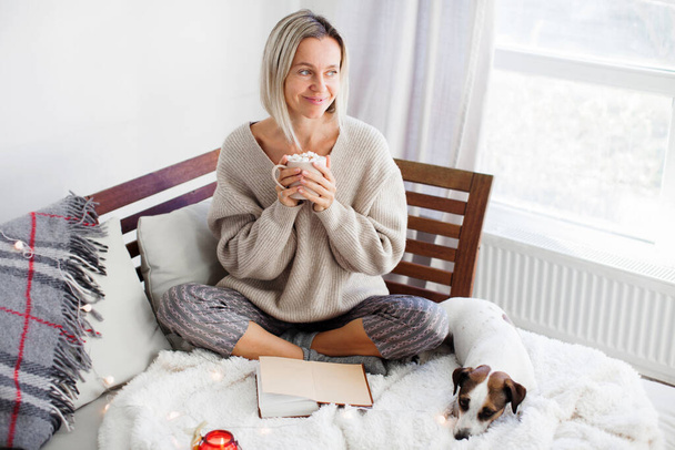 Adult woman sitting on the sofa at home and having a cup of coffee, looking away, middle aged lady with dog, relaxation concept - Photo, image