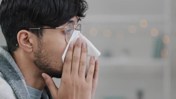 Close-up man gezicht zijaanzicht ziek Arabisch latino man bebaarde man met bril gewikkeld in deken lijdt aan loopneus niest allergie doekjes snot met papier servet respiratoire ziekte infectie - Video
