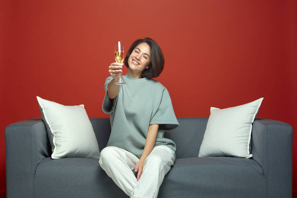 Joyful young girl raised glass of champagne smiling partying celebrating holiday event, sitting on sofa. Pajama party - Photo, Image