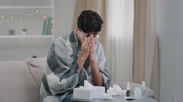 Arab bearded sick sad man with glasses sits on couch at home suffers from runny nose flu disease coronavirus pandemic covid epidemic sneezes. Unwell guy feeling bad fever virus illness symptoms indoor - Кадры, видео