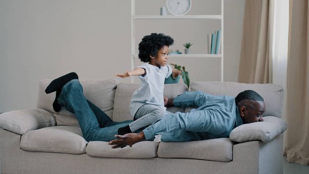 Adult african american father lying stomach on sofa little daughter plays sits on dads back kid girl pretends to fly on plane sitting on daddy spread arms to side mature man fun playing with child - 写真・画像