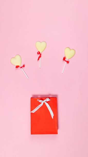 Καραμέλες λευκής σοκολάτας σε σχήμα καρδιάς κοντά σε κόκκινη τσάντα δώρου διακοσμημένη με λευκή κορδέλα. Έννοια των γλυκών δώρων για διακοπές. Επίπεδη. Αντιγραφή χώρου. Ανοιχτό ροζ φόντο. - Φωτογραφία, εικόνα