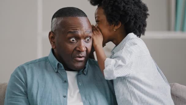 Little African American girl whispers in ear reveals secret to daddy daughter shares secrecy gossip confidential information mature father sitting on couch surprised from conversation opens eyes wide - Imágenes, Vídeo