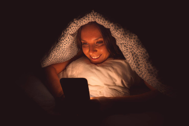 Happy smiling woman lying in bed under the blanket and typing on smartphone at night before sleeping. Social media addiction and communication - Photo, Image
