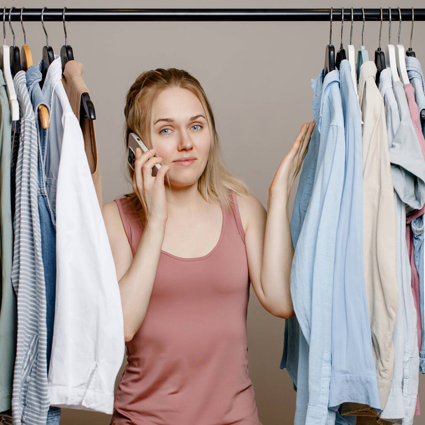 Pretty white woman shrugs her shoulders, doubting what to wear, Insecure woman standing near a clothes rack, not knowing what to wear to work. Nothing to wear and the concept of clothes.  - Foto, imagen