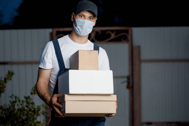 Male courier in the form of a delivery man in a medical mask holds a parcel in his hands in the open air. Concept of courier at any time will deliver your parcel.  - Photo, Image