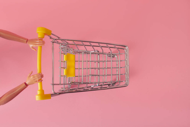 Кукольные руки толкают тележку из супермаркета на розовый фон, концепция минимализма - Фото, изображение