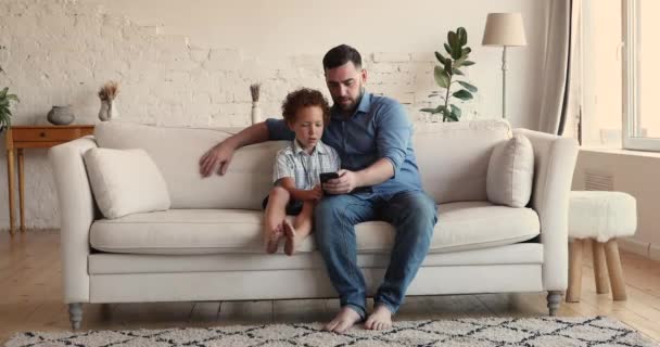 Dad and 5s son sit on couch using smart phone - Materiał filmowy, wideo