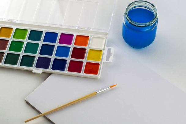 watercolor paints a brush for drawing a jar of water and a sketchbook for drawing on a white surface. High quality photo - Foto, afbeelding