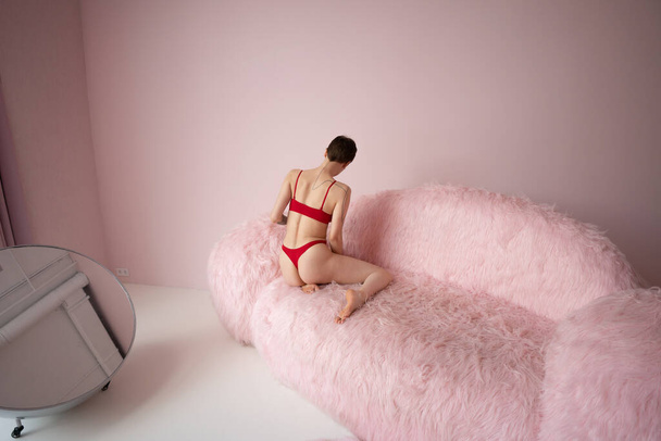 молода красива жінка позує в червоній білизні в студії, сидячи на рожевому дивані
 - Фото, зображення