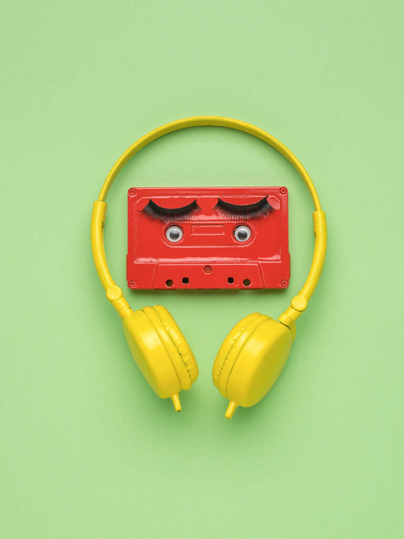 A red tape recorder with eyes and yellow headphones on a light green background. Creative image. The concept of analog sound recording. Flat lay. - Фото, зображення
