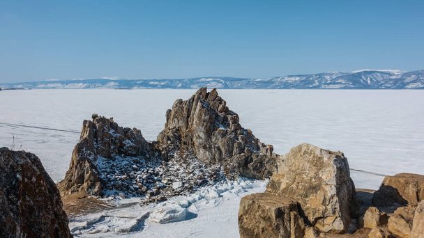 A picturesque granite double-headed rock on the background of a frozen lake. Cracks on slopes devoid of vegetation. A mountain range in the distance. Boulders in the foreground. Blue sky. Baikal - Photo, Image
