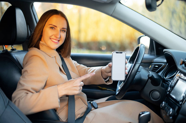 Smiling woman sits in the car and showing smartphone with empty blank screen - Fotó, kép