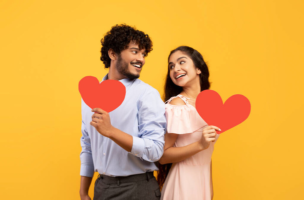 Valentines holiday. Portrait of indian lovers holding red paper hearts and looking at each other, yellow background - 写真・画像