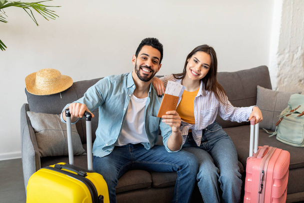 New trip, finally. Excited young tourists couple holding tickets and passports, sitting with packed suitcases on sofa - Foto, imagen