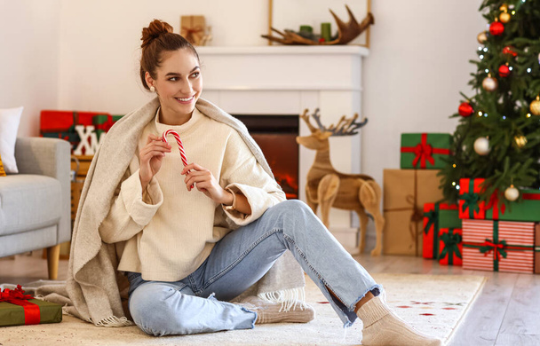 Pretty young woman with tasty candy cane at home on Christmas eve - Photo, image