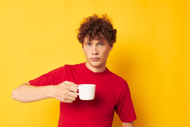 Young curly-haired man in a red T-shirt with a white cap in his hands isolated background unaltered - Фото, зображення