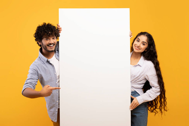 Happy indian couple in love standing by white empty board for advertisement or text over yellow studio background - Foto, immagini
