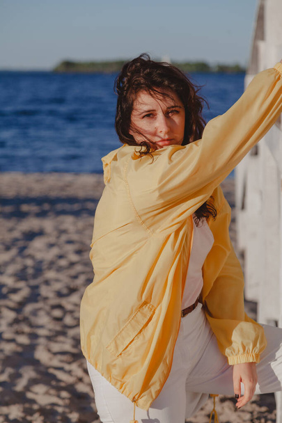 Szép lány sárga dzsekiben nyugszik egy fehér fa szerkezeten a homokos parton. - Fotó, kép